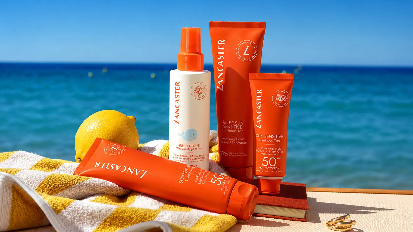 Tube de crème solaire Orange Lancaster SPF 50+ sensible au soleil, avec un flacon pulvérisateur blanc et un capuchon orange assorti à côté, reposant sur une serviette rayée jaune et blanche près de l’océan.