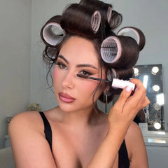Une femme aux cheveux foncés coiffés avec plusieurs bigoudis en plastique transparents applique du mascara sur ses cils supérieurs.