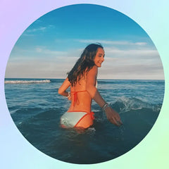 Une femme en bikini blanc et orange avec des liens rouges se tient dans l’océan jusqu’à la taille, souriante alors qu’elle tend la main vers de douces vagues.