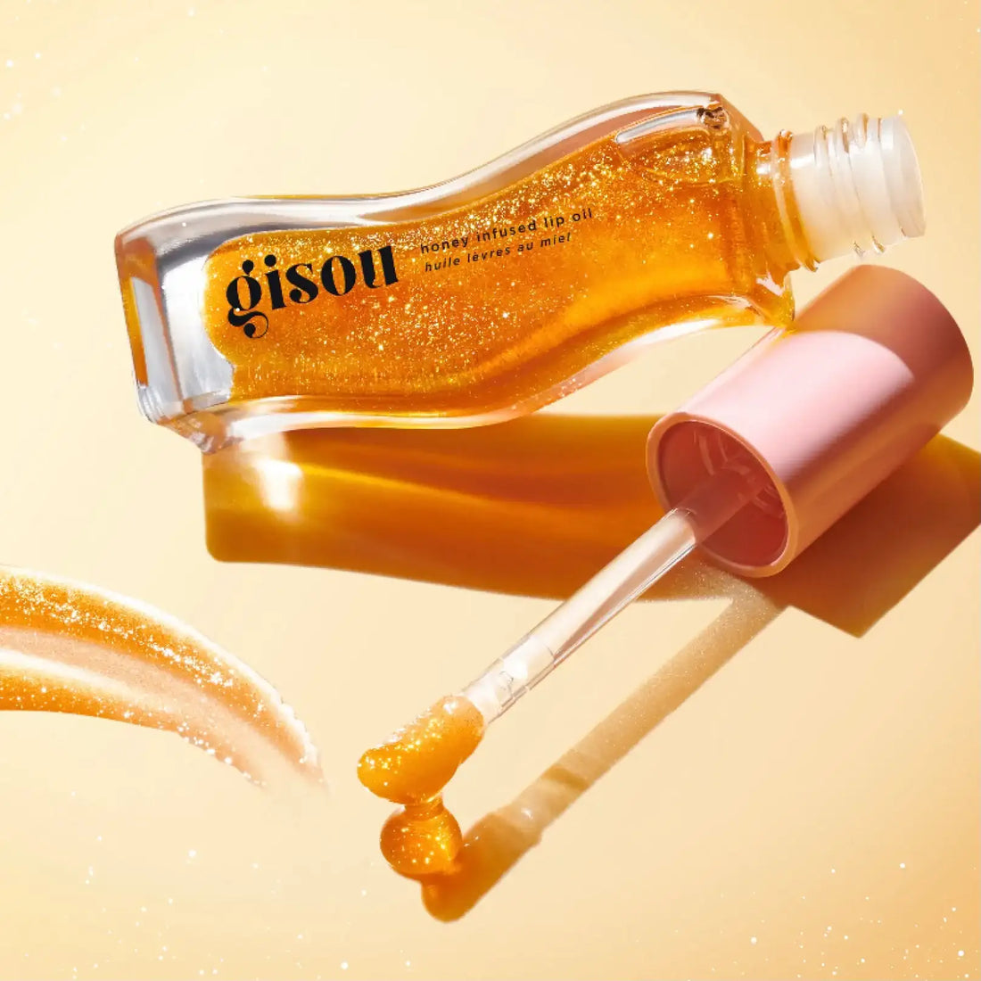 Gisou : huile pour les lèvres infusée au miel, dans un flacon en verre transparent avec un bouchon rose et un liquide doré scintillant.
